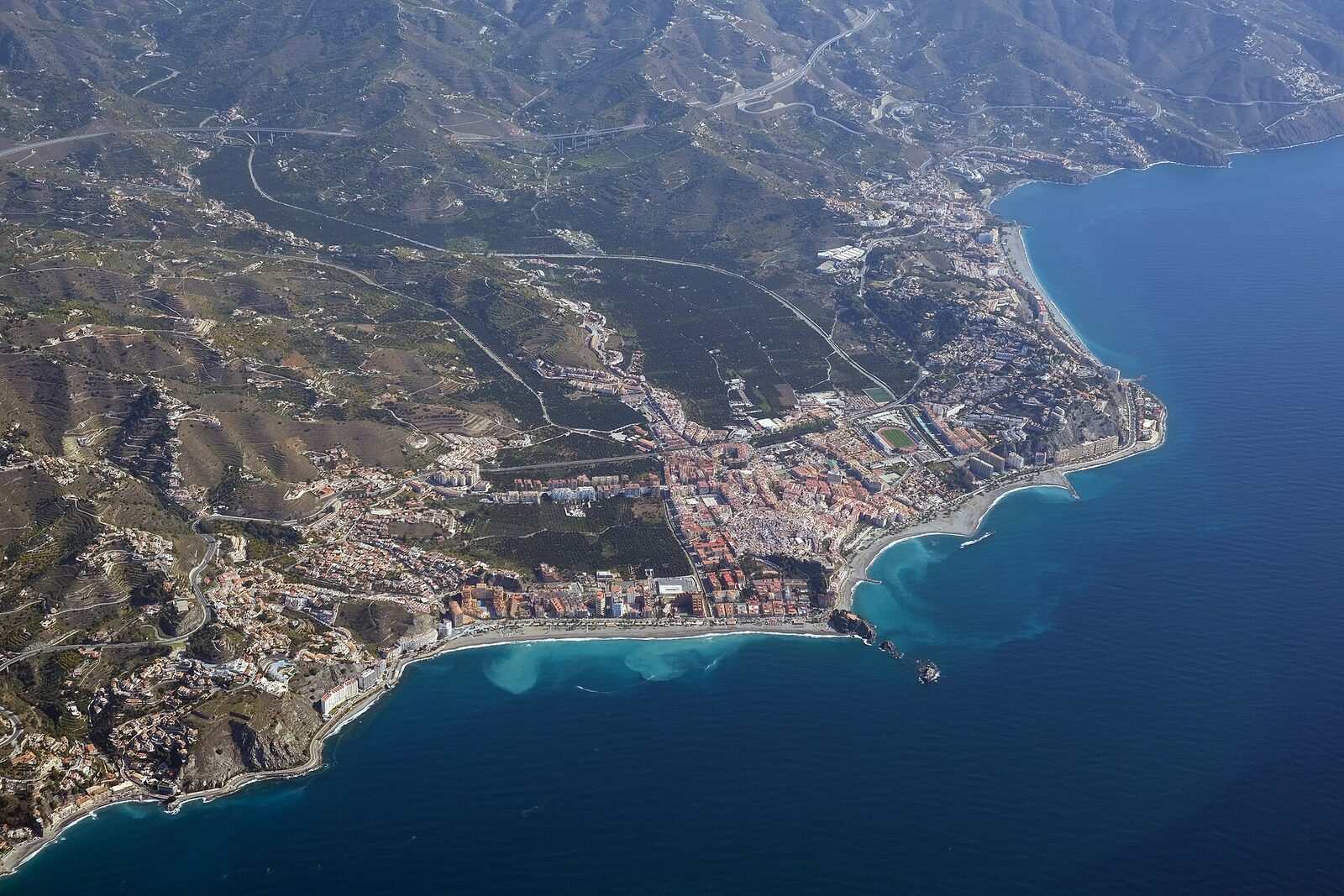 Almuñécar från luften: Peñón del Santo delar stranden i två, med Puerta del Mar till vänster och San Cristóbal till höger.